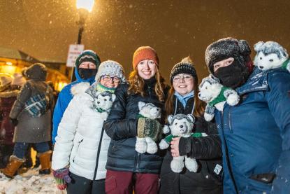 five people wearing winter attire smiling and holding husky stuffed animal toys 